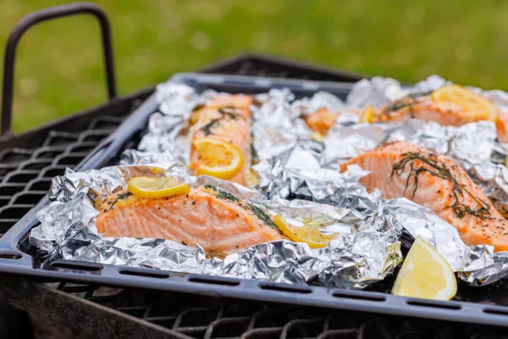 Salmon fillets with lemon slices and herbs are wrapped in foil and cooked on a grill outside.