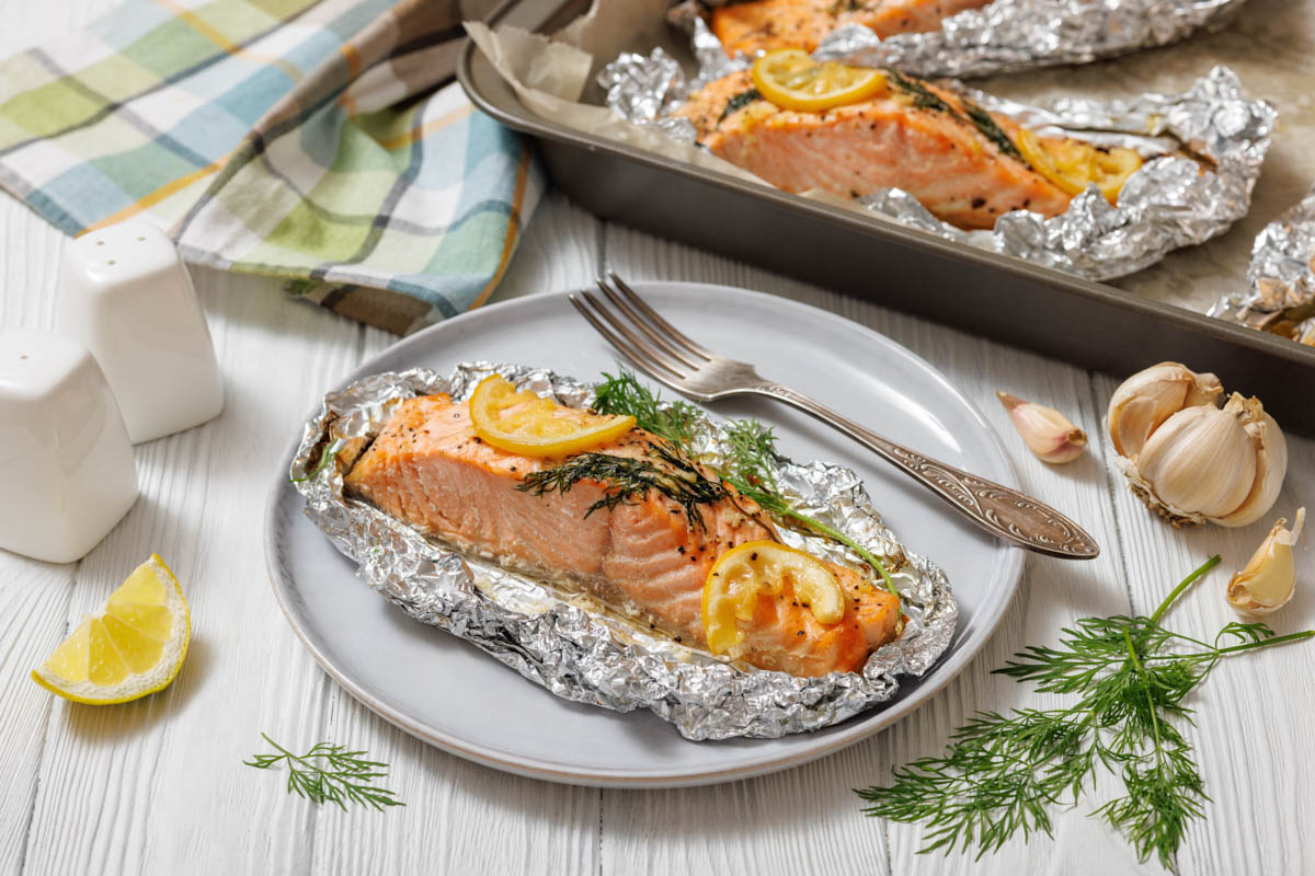 A close-up, slightly high-angle shot of a single Lemon Dill Salmon Foil Packets , seasoned with lemon and dill, still nestled in its aluminum foil packet on a light gray plate, with a silver fork to its right.
