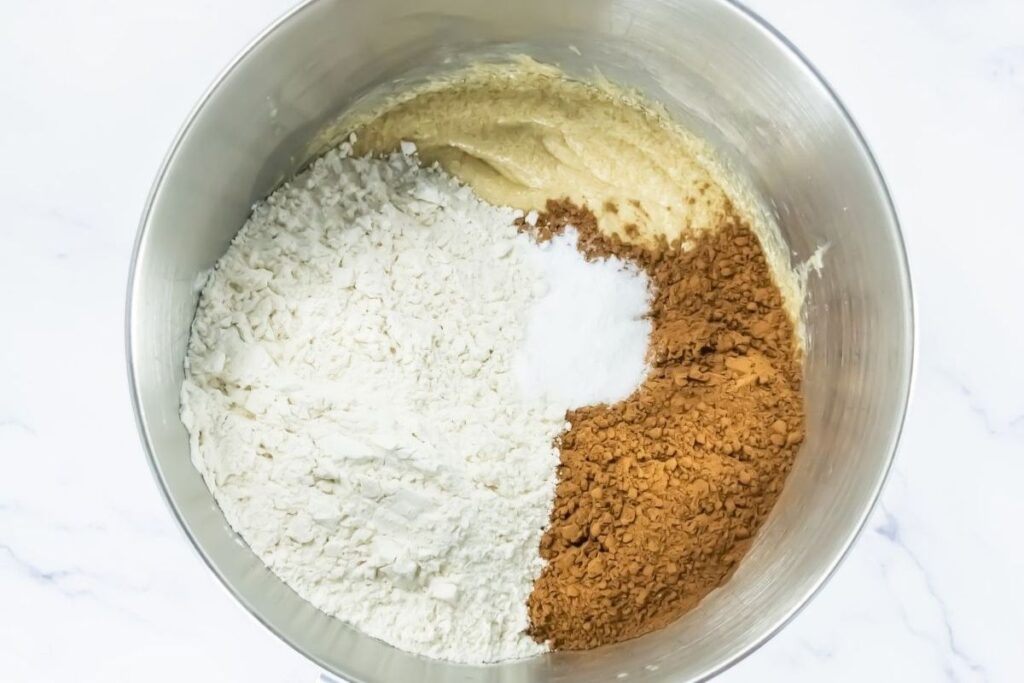 A mixing bowl containing flour, baking soda, cocoa powder, and creamed butter and sugar, ready to be combined.