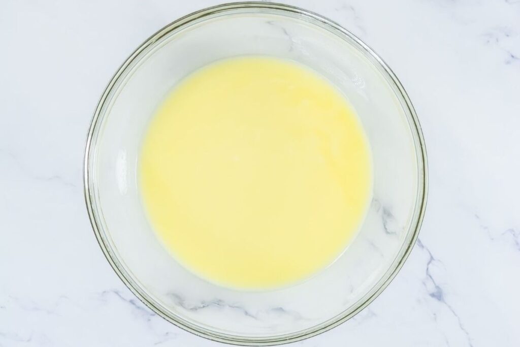 A clear glass bowl containing a pale yellow liquid mixture sits on a white marble surface.
