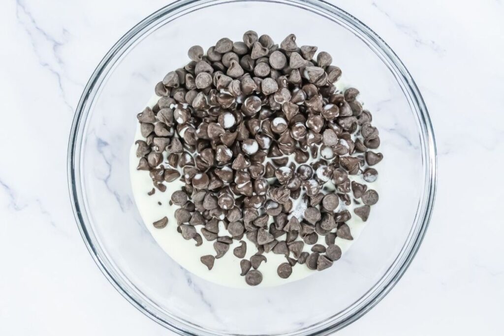 A clear glass bowl containing chocolate chips on top of a layer of cream, placed on a white marble surface.