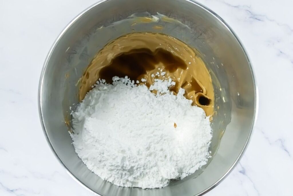 A metal mixing bowl containing peanut butter and powdered sugar on a white marble surface.