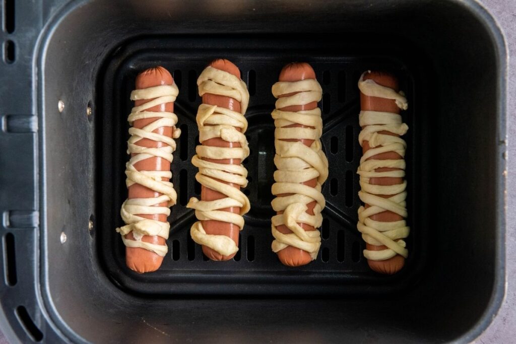Four hot dogs wrapped in strips of dough are placed inside an air fryer basket, ready to be cooked.
