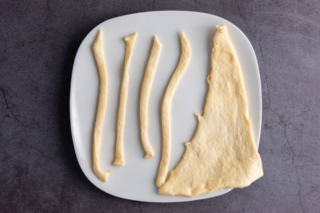 A white plate holds several strips of raw dough and one larger, irregularly shaped piece of dough, set on a dark gray surface.
