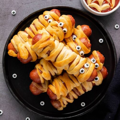 A plate of hot dogs wrapped in dough to resemble mummies, with candy eyes, next to a bowl of ketchup decorated with a spiderweb pattern.