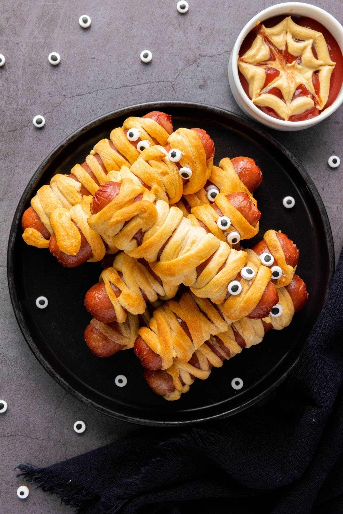 A plate of hot dogs wrapped in dough to resemble mummies, with candy eyes, next to a bowl of ketchup decorated with a spiderweb pattern.