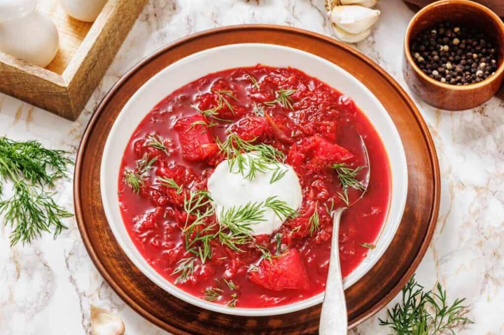A bowl of borscht with a dollop of sour cream and fresh dill on top, served on a brown plate with a spoon on a marble surface.