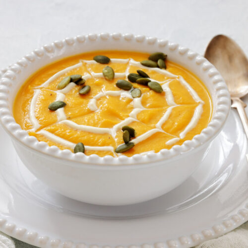 A white bowl of pumpkin soup topped with a swirl of cream and pumpkin seeds, placed on a white plate with a spoon beside it.
