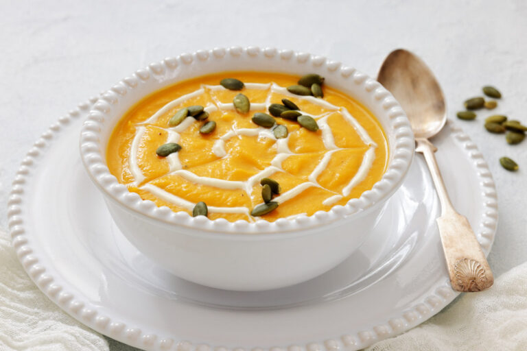 A white bowl of pumpkin soup topped with a swirl of cream and pumpkin seeds, placed on a white plate with a spoon beside it.