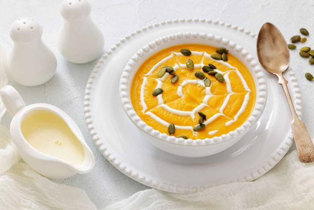A bowl of creamy pumpkin soup garnished with cream and pumpkin seeds, served with a spoon, a small pitcher of cream, and salt and pepper shakers on a white surface.