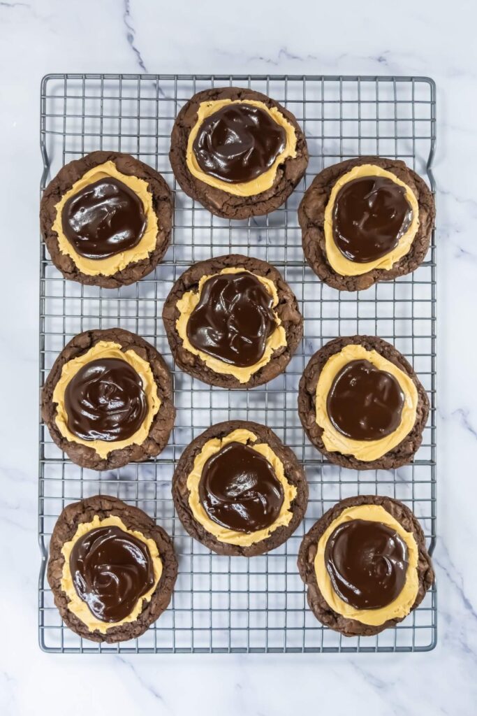 Nine chocolate cookies with a layer of peanut butter and a glossy chocolate topping are arranged on a cooling rack over a marble surface.