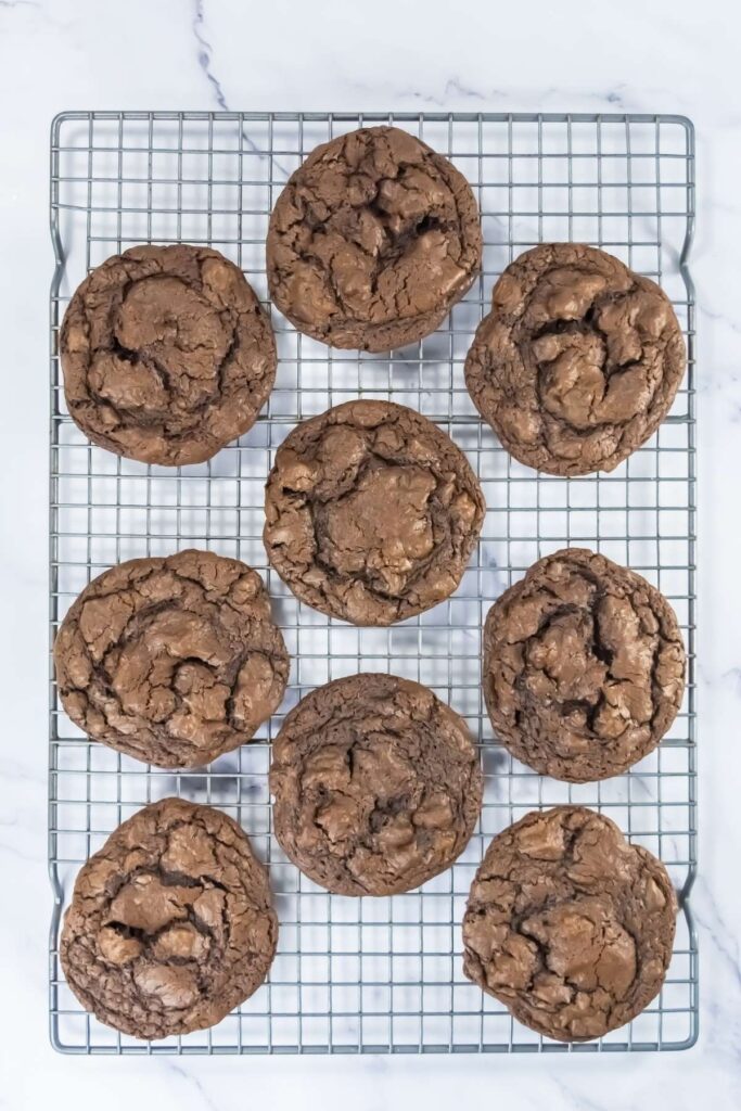 Nine chocolate cookies with a cracked surface cool on a metal wire rack set on a white marble background.
