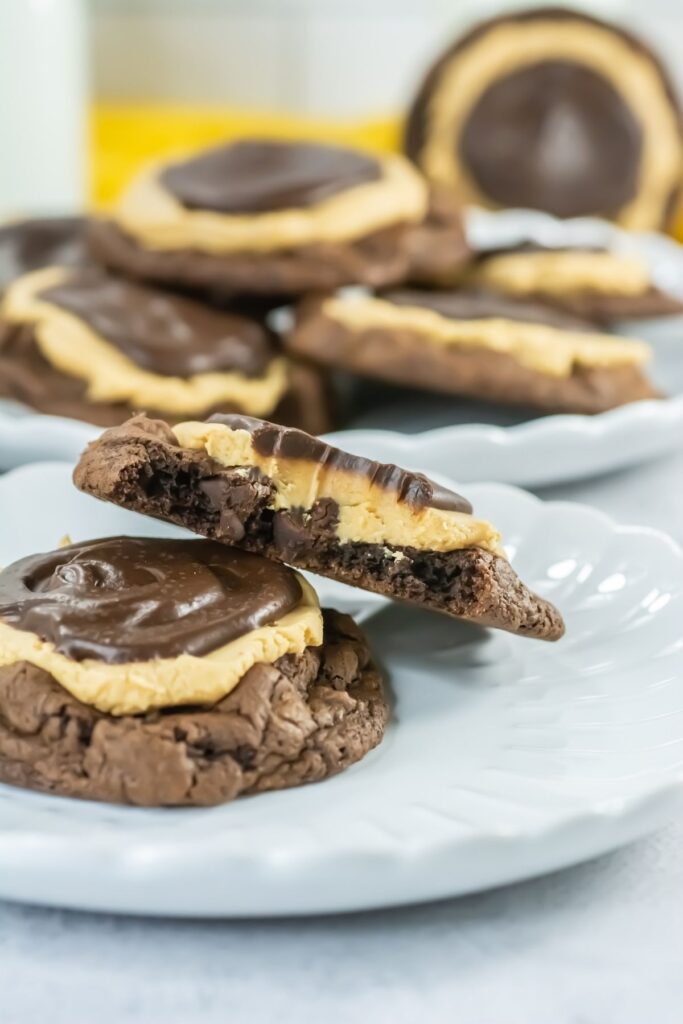 A chocolate cookie topped with peanut butter and a chocolate layer, cut in half on a white plate, with more cookies in the background.