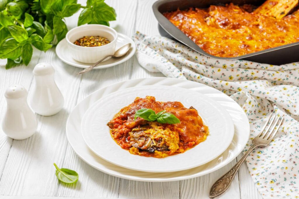 A plate of baked eggplant parmesan topped with basil, served with a fork; casserole dish, herbs, spices, and salt and pepper shakers in the background.