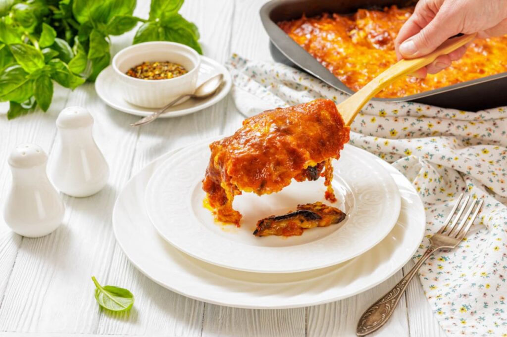 A hand serves a slice of baked lasagna with tomato sauce onto a white plate, with a baking dish, fresh basil, and condiments in the background.