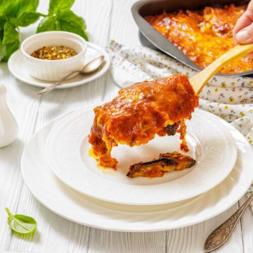 A hand serves a slice of Eggplant Parmigiana onto a white plate, with a casserole dish, herbs, and condiments visible on a white table.