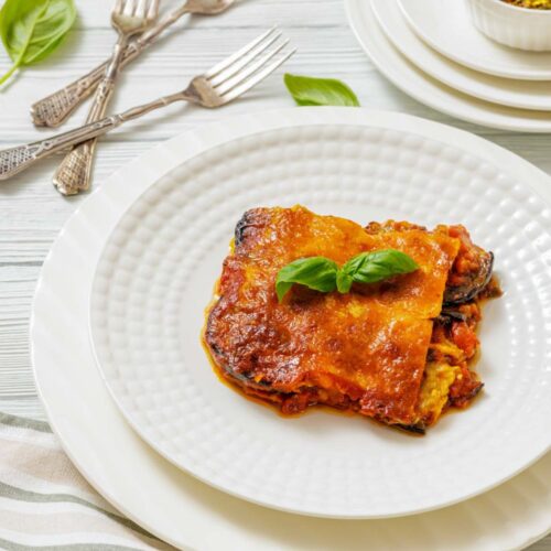 A slice of baked lasagna topped with basil is served on a white plate, with stacked dishes, cutlery, and a small bowl of spices in the background.