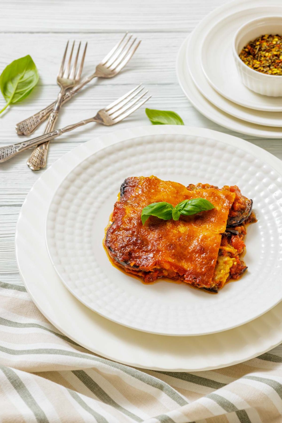 A slice of baked lasagna topped with basil is served on a white plate, with stacked dishes, cutlery, and a small bowl of spices in the background.