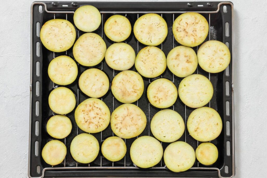 Sliced eggplants arranged in a single layer on a black baking tray with a metal rack.