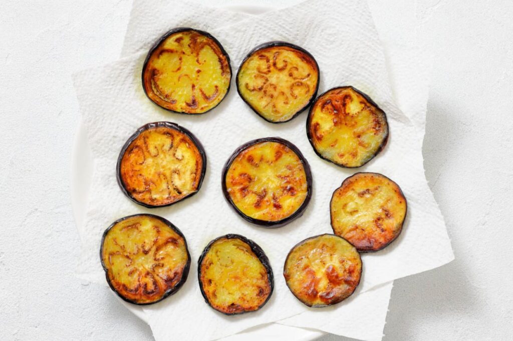Round slices of roasted eggplant arranged on a paper towel atop a white plate, showing browned tops and a light, textured background.