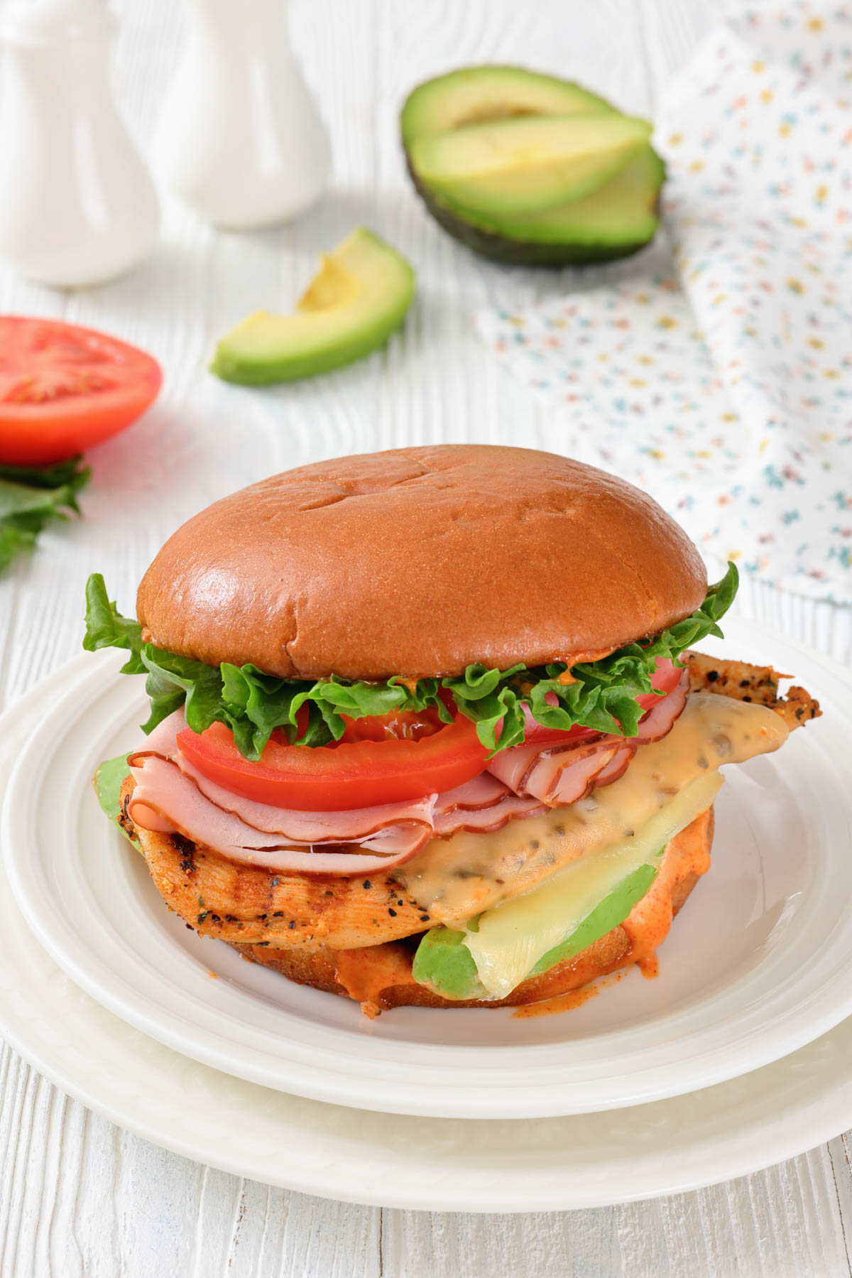 A chicken sandwich with lettuce, tomato, sliced ham, cheese, and avocado on a bun is plated, with avocado, tomato, and condiments in the background.