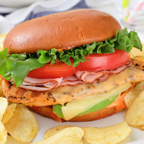 A grilled chicken sandwich with lettuce, tomato, cheese, and sliced ham on a bun, served with potato chips on a white plate.