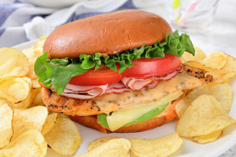 A grilled chicken sandwich with lettuce, tomato, cheese, and sliced ham on a bun, served with potato chips on a white plate.