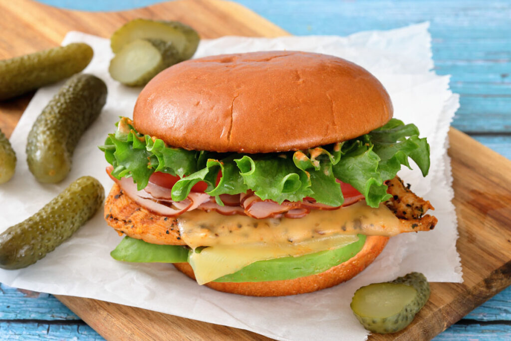A grilled chicken sandwich with lettuce, tomato, cheese, onions, and avocado on a bun, served with pickles on a wooden board.