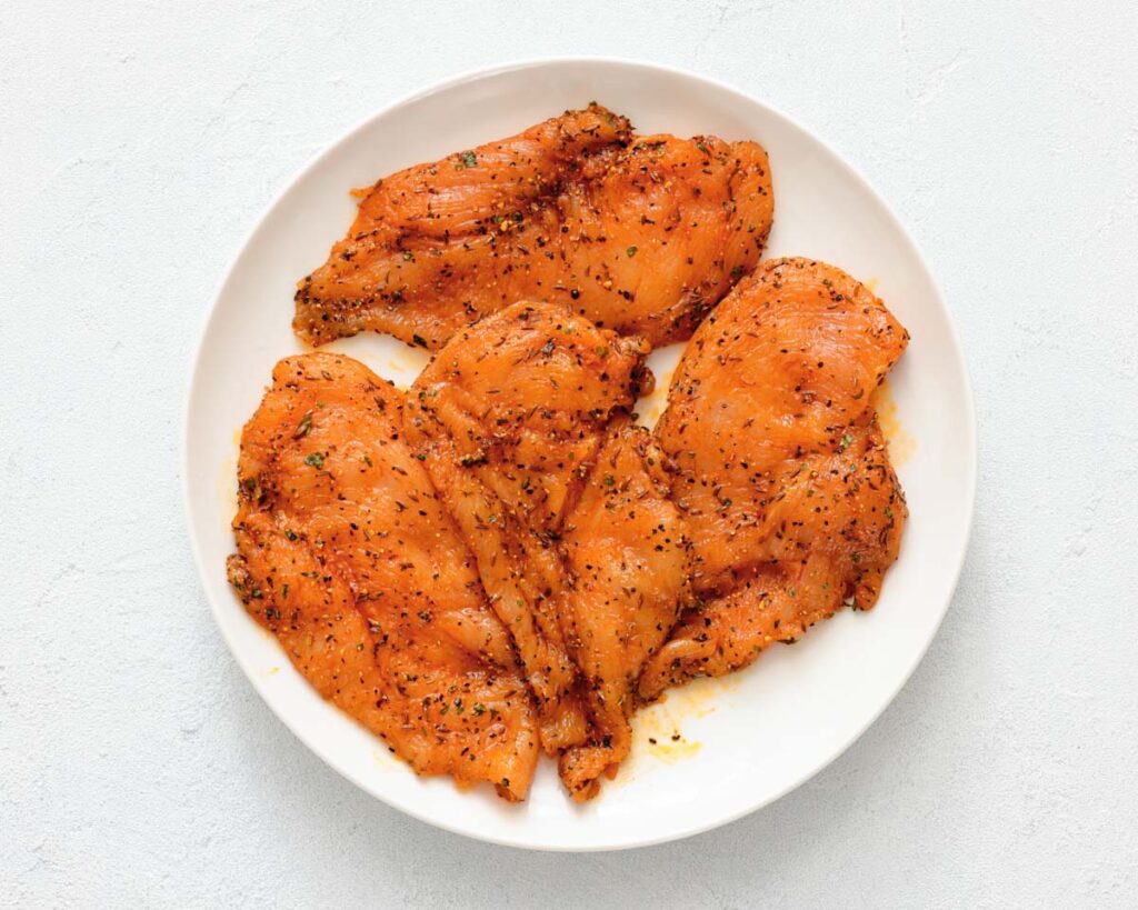 Four raw, seasoned chicken breasts arranged on a white plate against a white background.