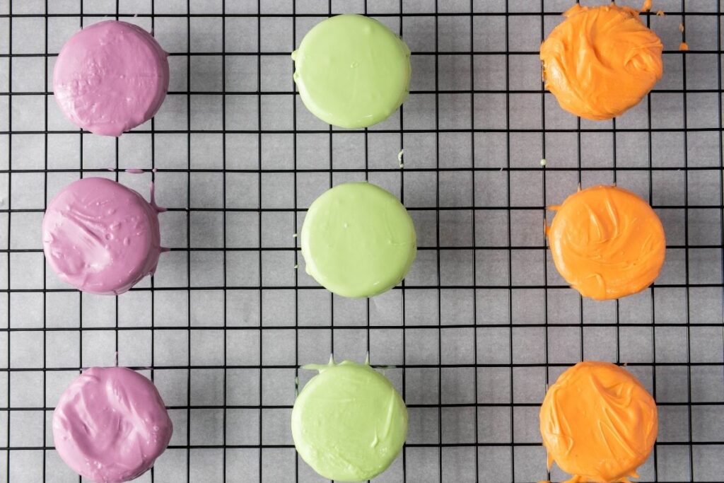Nine round cookies with smooth purple, green, and orange coating arranged in rows on a black wire cooling rack.