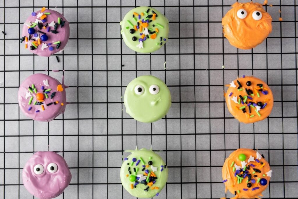Nine decorated cookies with colorful coating and sprinkles, some with candy eyes, arranged in a grid pattern on a cooling rack.