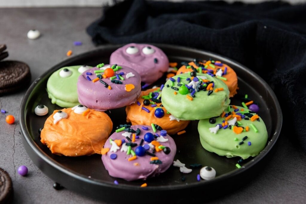 A black plate with cookies coated in purple, orange, and green, topped with sprinkles and candy eyes, on a gray surface.
