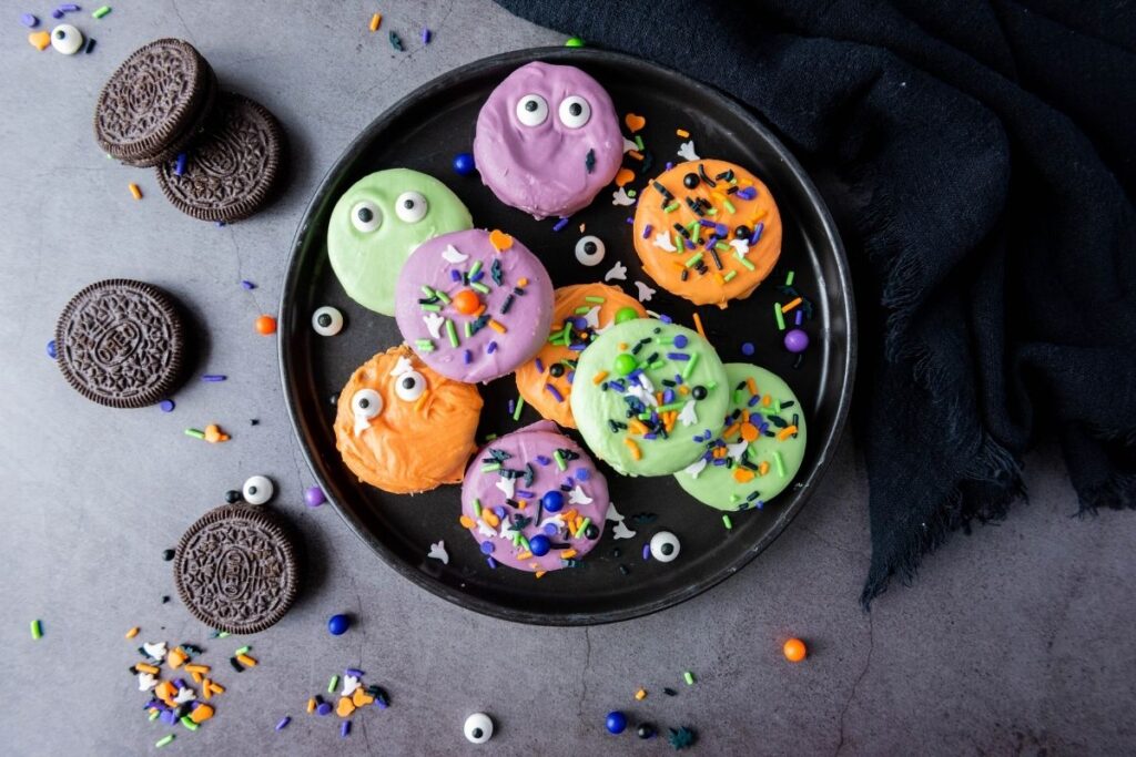 A black plate with colorful, coated cookies decorated with candy eyes and sprinkles, surrounded by Oreos and scattered decorations on a gray surface.
