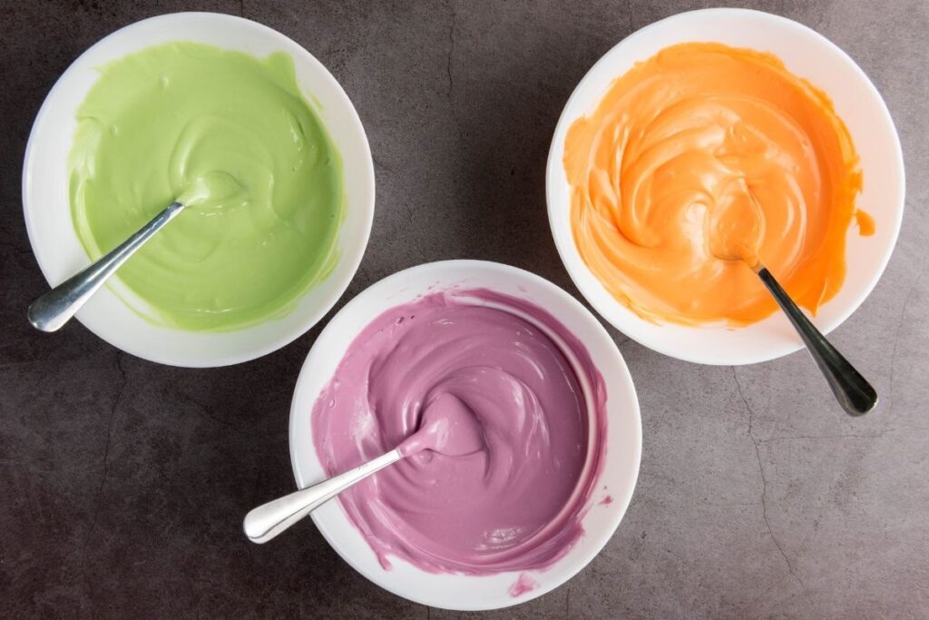 Three white bowls, each with a metal spoon, contain pastel green, orange, and purple melted candy on a gray surface.