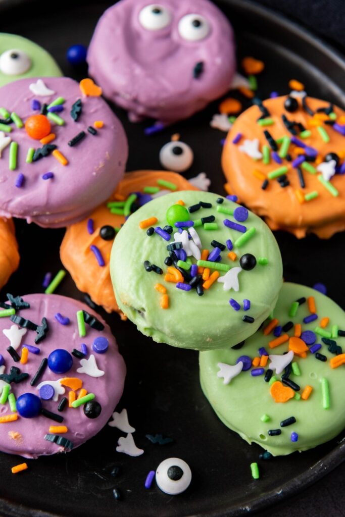 Colorful, coated cookies decorated with Halloween-themed sprinkles and candy eyes, arranged on a black plate.