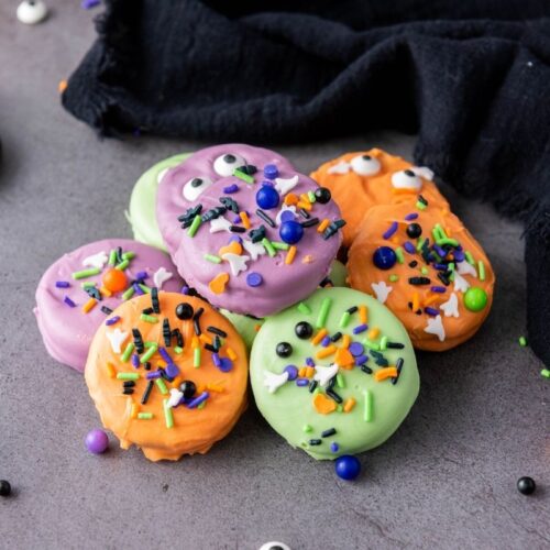 A group of Halloween Oreo Cookies coated in green, orange, and purple, decorated with sprinkles and candy eyes, sits on a gray surface next to a black cloth.