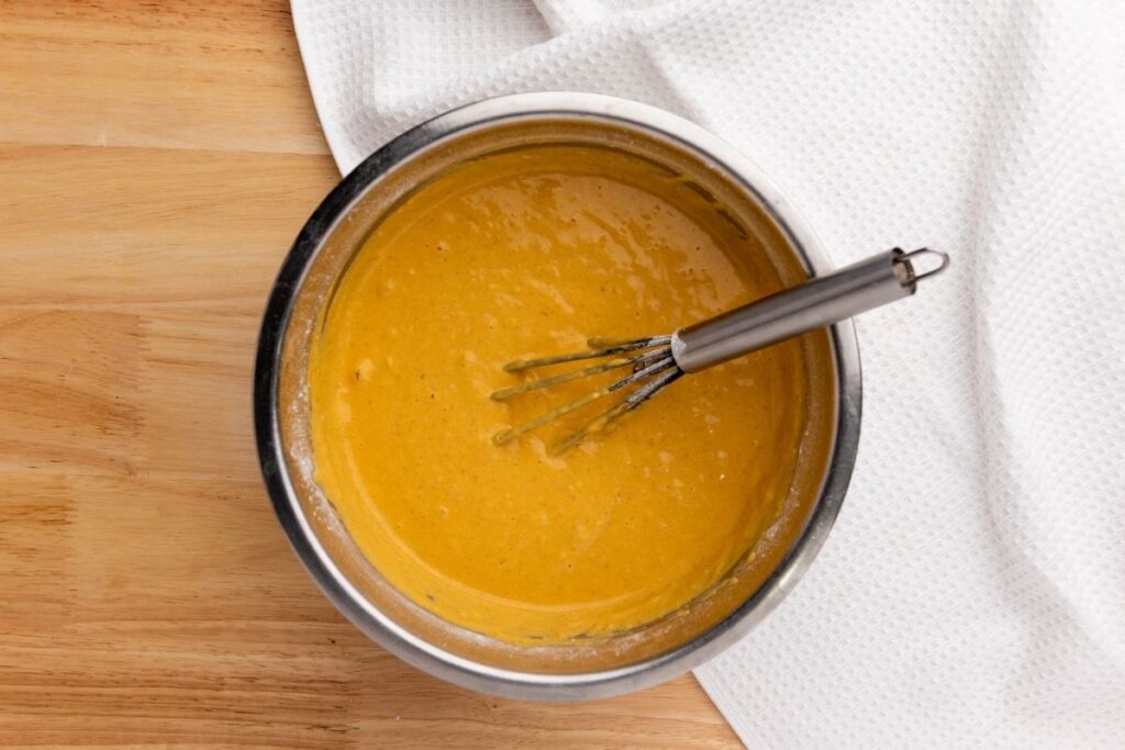 A metal bowl with thick orange batter and a whisk inside, placed on a wooden surface next to a white textured cloth.