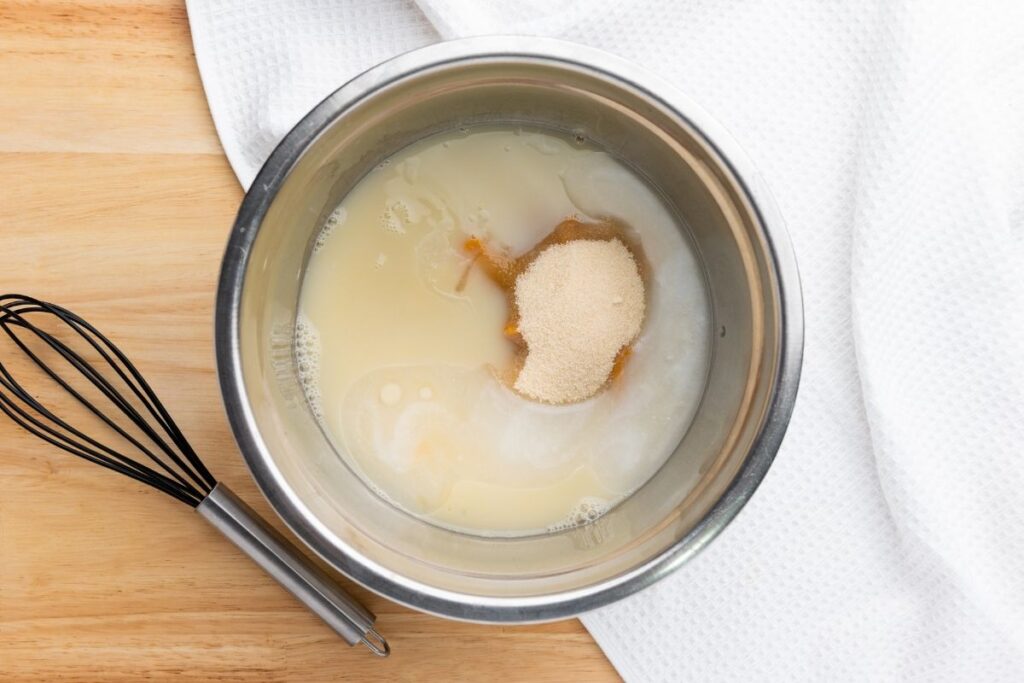 A metal mixing bowl with pumpkin puree, sugar, and liquid ingredients next to a whisk on a wooden surface with a white towel.