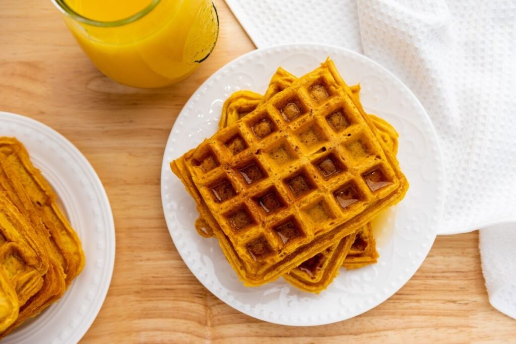 A white plate with stacked waffles sits on a wooden table next to a glass of orange juice and a white textured cloth.