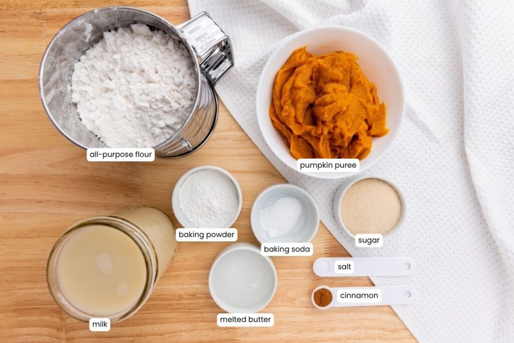 A wooden surface displays bowls of all-purpose flour, pumpkin puree, baking powder, baking soda, sugar, milk, melted butter, salt, and cinnamon, each labeled.