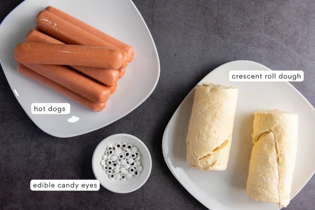 A plate of hot dogs, a plate with two rolls of crescent roll dough, and a bowl of edible candy eyes are shown on a dark surface.