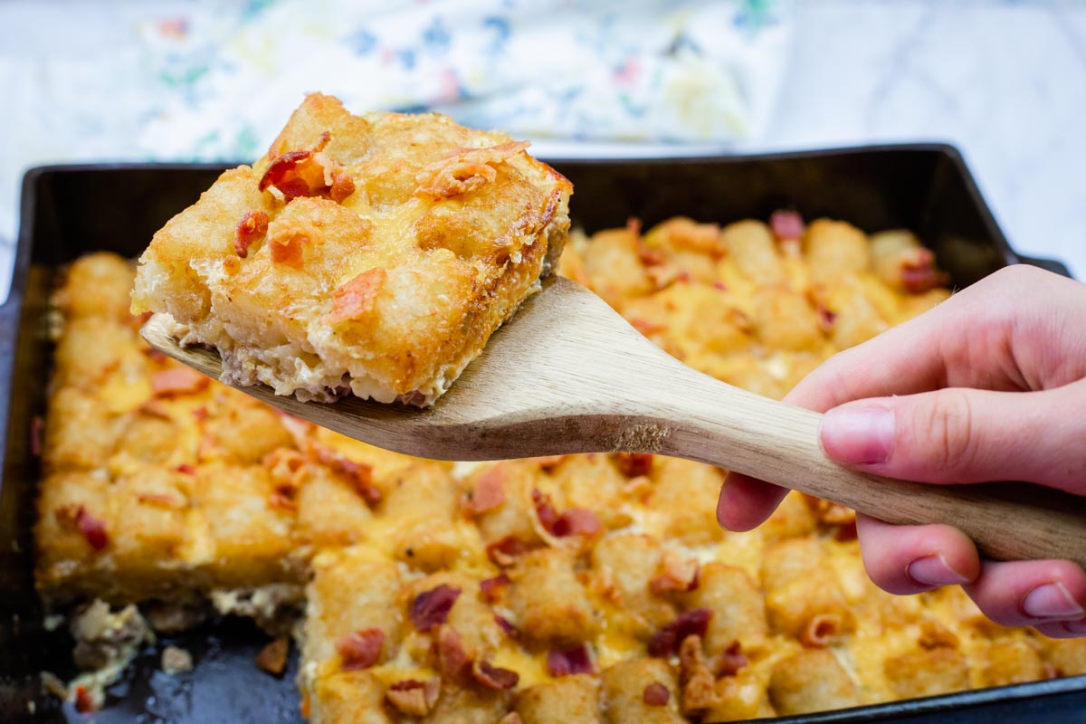 A hand holds a wooden spatula lifting a Cheesy Tater Tot Breakfast Casserole from a baking tray.