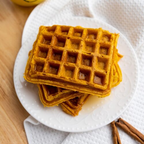 A plate with three stacked waffles sits on a white cloth with two cinnamon sticks nearby and a glass of orange juice in the background.