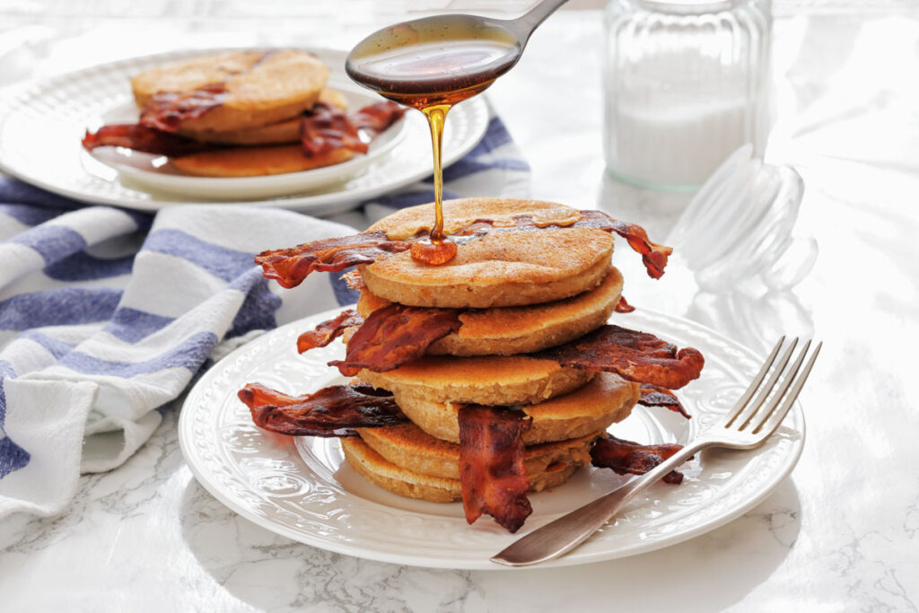 A stack of pancakes with crispy bacon is being drizzled with syrup, set on a white plate with a fork, with milk and another plate in the background.