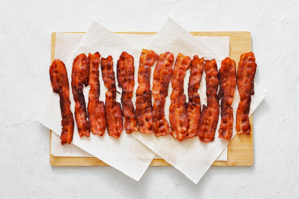 Crispy cooked bacon strips are arranged in a row on paper towels atop a wooden cutting board.