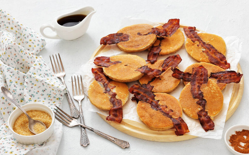 A round wooden tray with pancakes each topped with a strip of bacon, served alongside syrup, brown sugar, and four forks on a white table.
