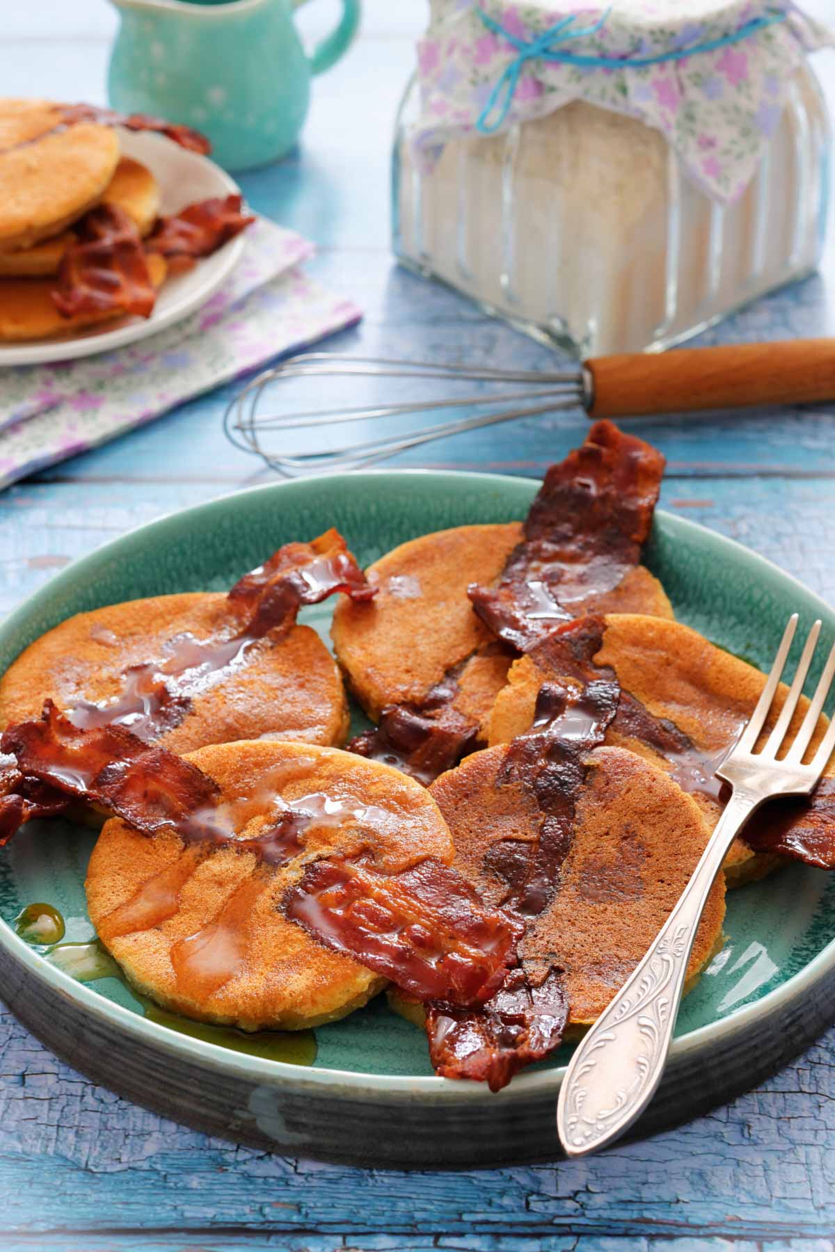 A plate of Bacon Pancakes and syrup, with a fork on the side; a whisk, jar, and plate with more pancakes and bacon in the background.