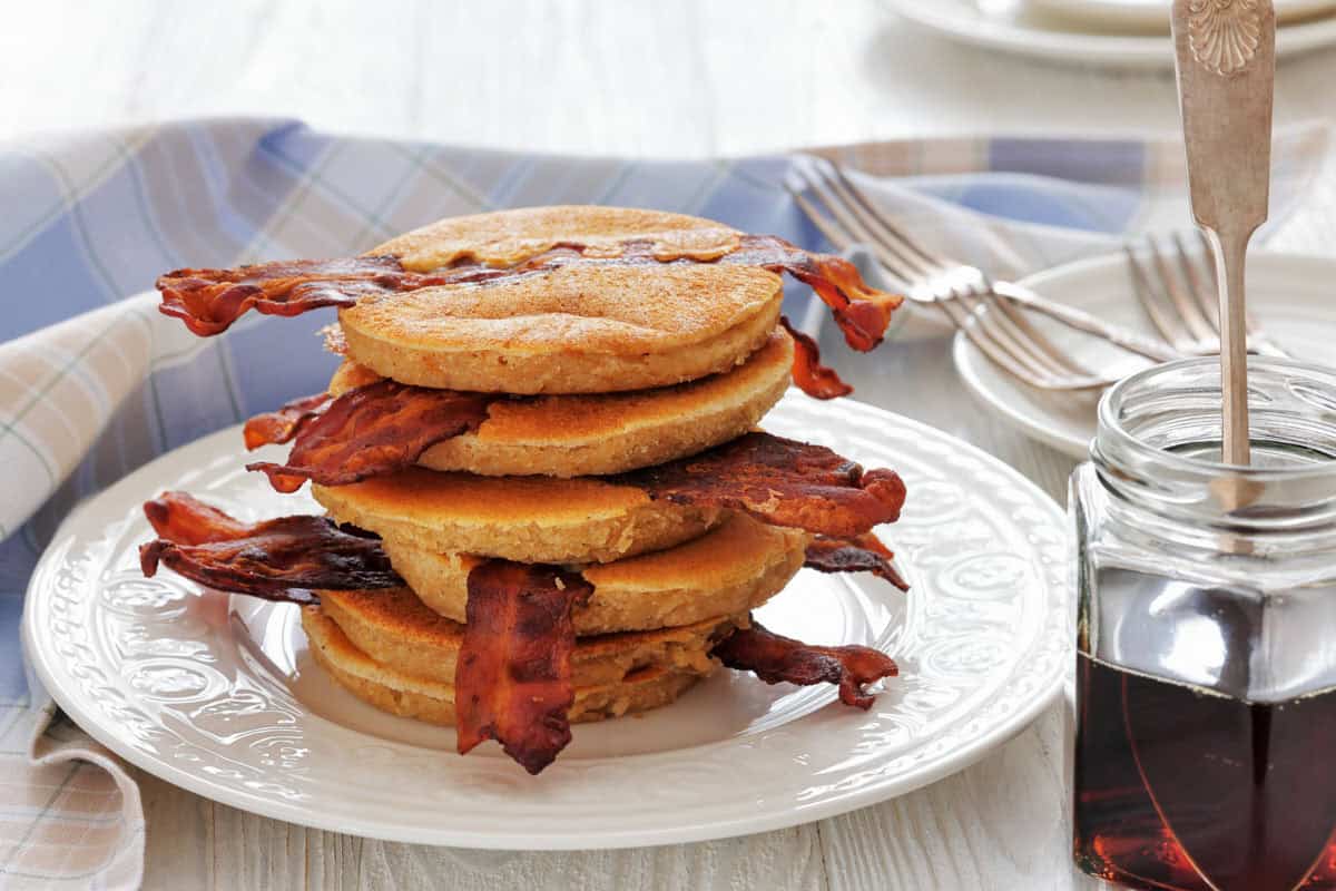 A stack of pancakes with strips of bacon between layers sits on a white plate, next to a jar of syrup and a folded napkin with forks.