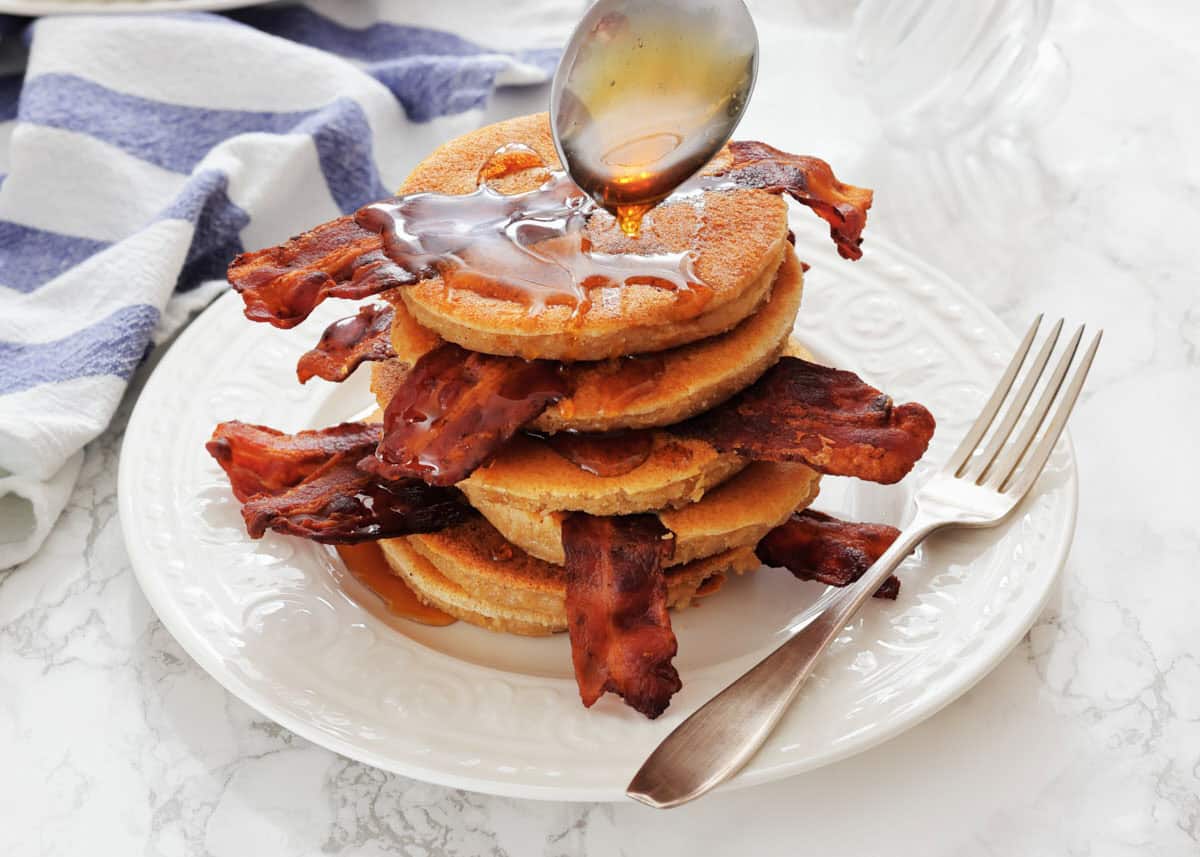 A stack of pancakes with strips of bacon, being drizzled with syrup from a spoon, on a white plate with a fork beside it.