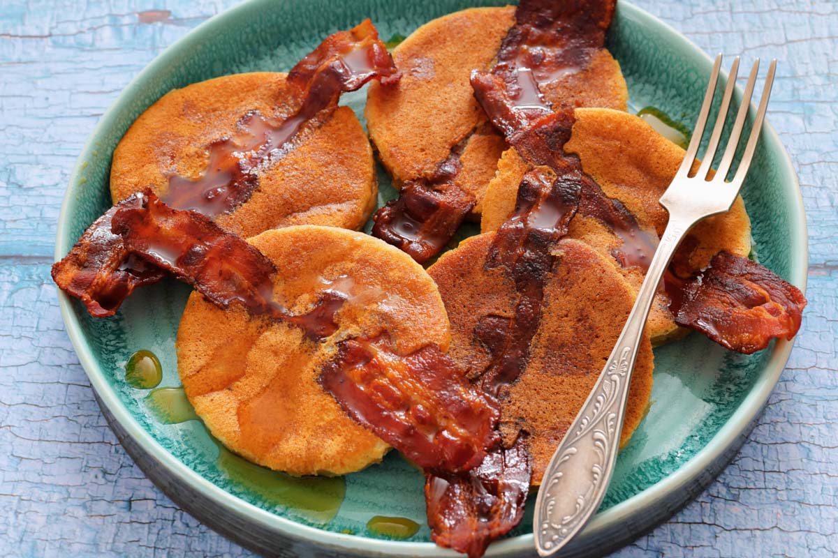 A plate with five Bacon Pancakes and drizzled syrup, with a fork resting on the side.